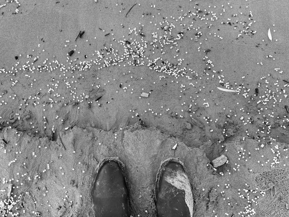 photograph of a persons boots with microplastics littering the beach around them.